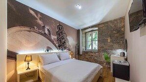A bedroom with a bed, a nightstand with a lamp, a stone wall, a window, and a small desk with a kettle. - Pjaca City Rooms (Split)