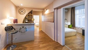 A modern hotel room with a wooden floor, a bar area with a decorative emblem, a clock, and a bed with a green chair. - Pjaca City Rooms (Split)