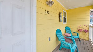 A yellow house with the number 105 on the wall, a porch with turquoise Adirondack chairs, and a small pink table. - Dream Chaser- Walk to Sandfest & Ocean Breeze! Heated Pool & Pets OK (Port Aransas)
