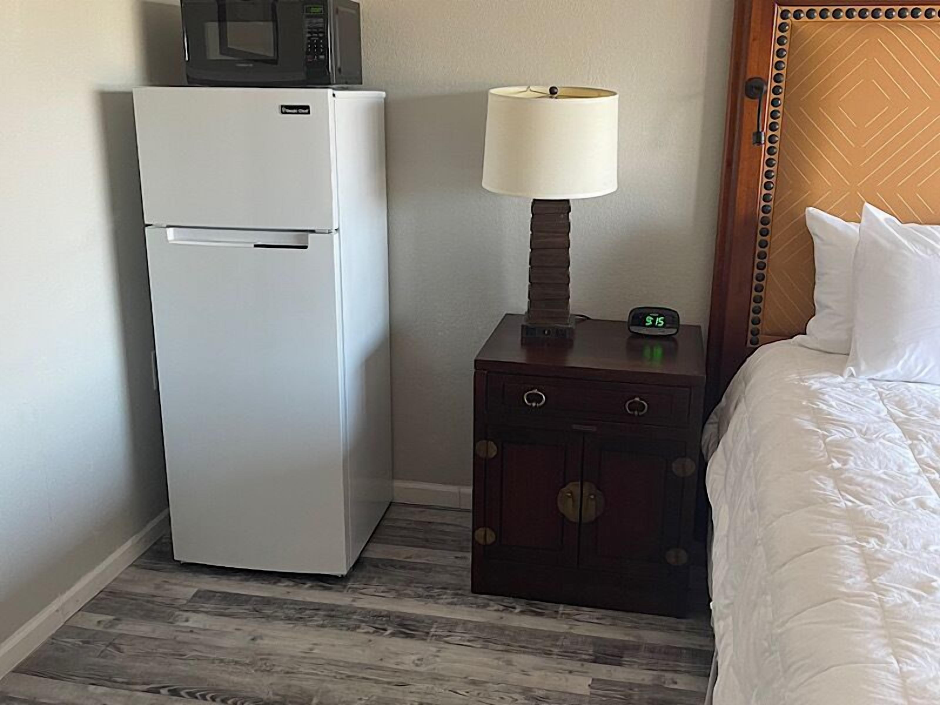 Une chambre d’hôtel de taille modeste, dotée d’un réfrigérateur blanc, d’une lampe de chevet, d’une horloge et d’un lit avec une literie blanche.