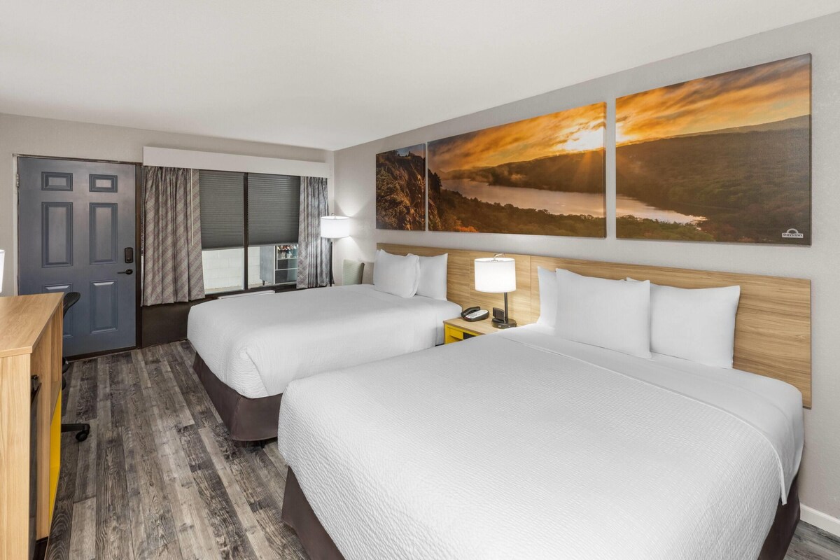 Habitación de hotel con dos camas, un escritorio, una silla, una lámpara y un ventanal grande con cortinas.