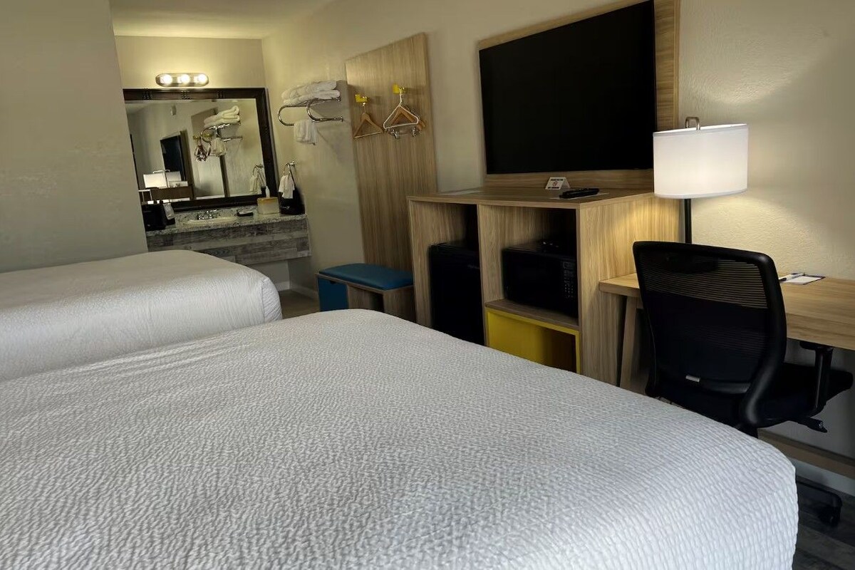 A hotel room with two beds, a desk with a chair, a TV, a lamp, and a mirror.