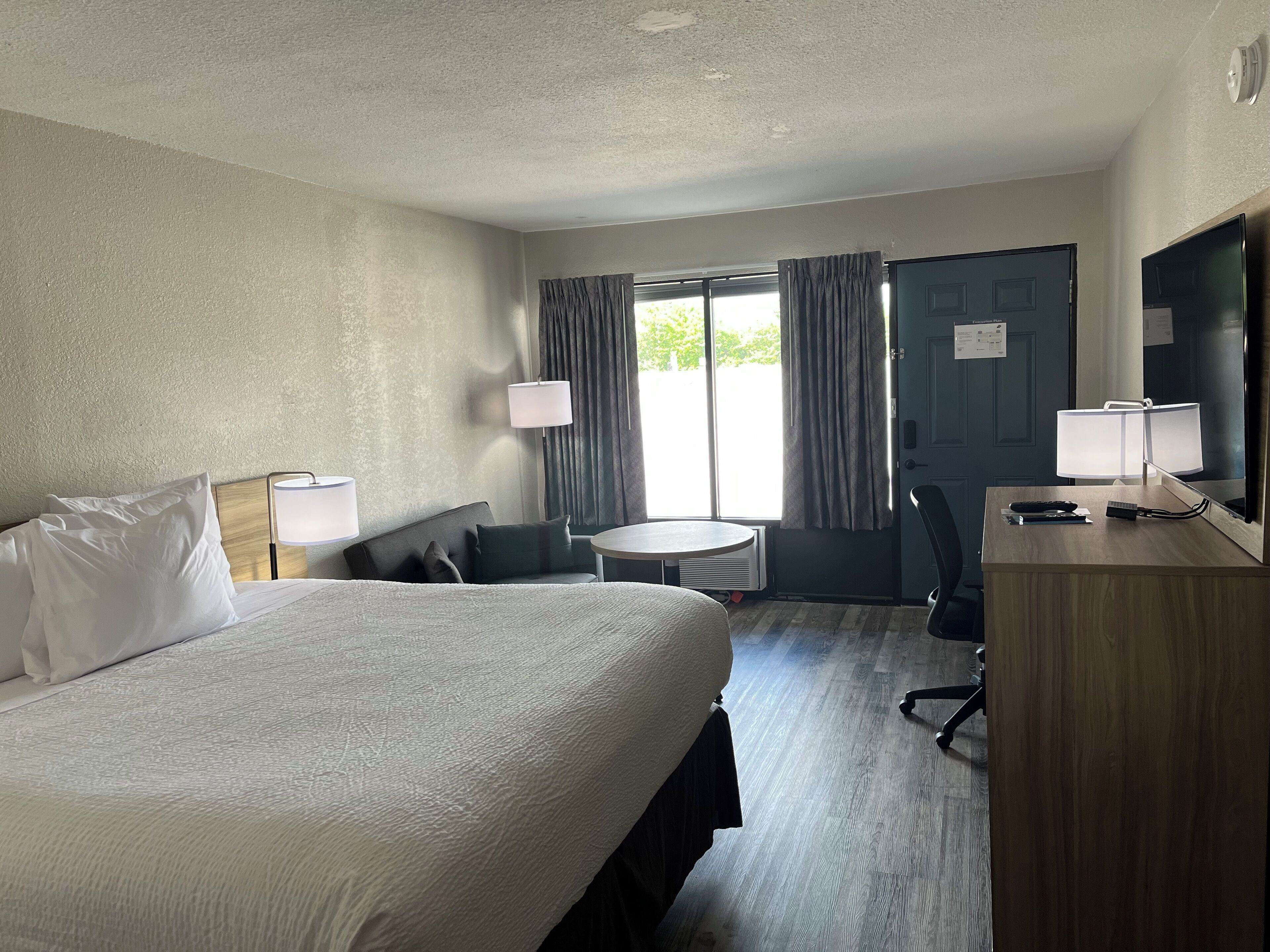 A hotel room with a bed, a desk with a lamp, a chair, a small table, and a TV.
