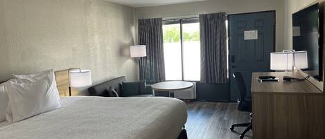 A hotel room with a bed, a desk with a lamp, a chair, a small table, and a TV.