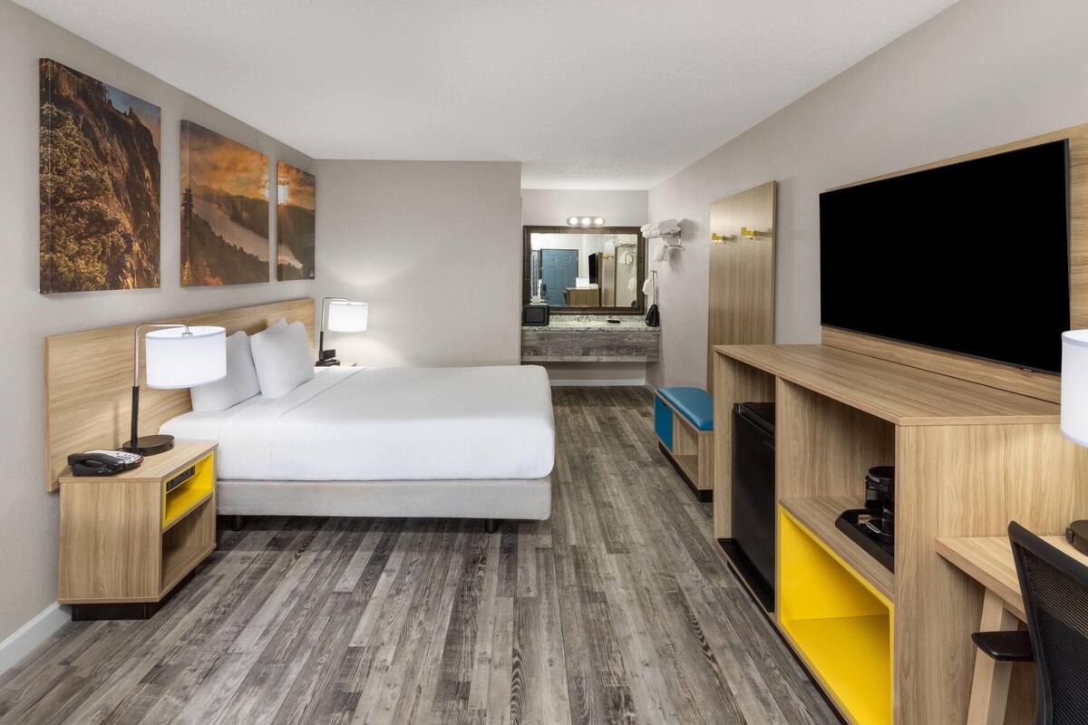 A hotel room with a bed, bedside table, lamp, TV, and a desk with a chair.