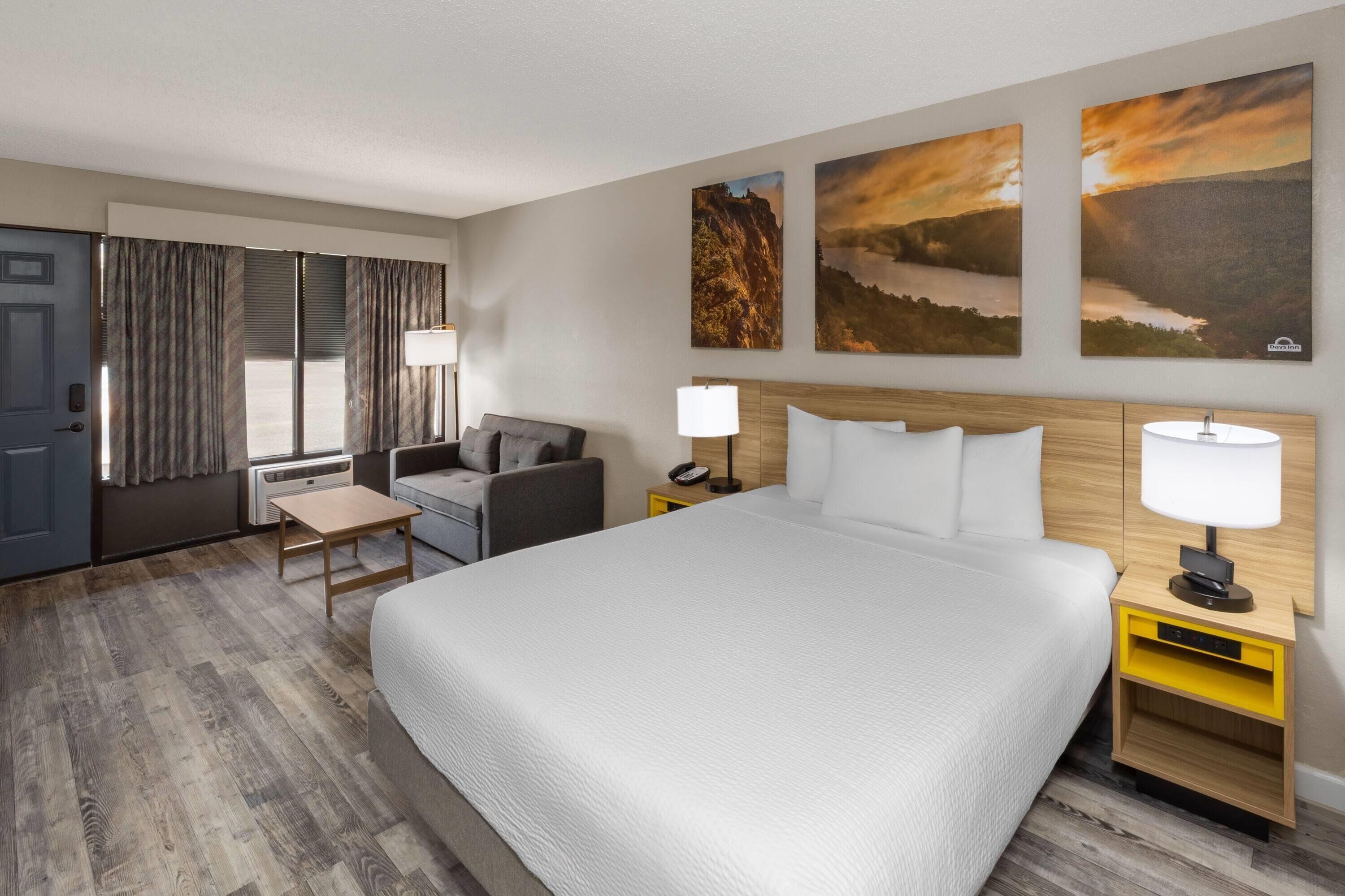 A hotel room with a large bed, a desk, a chair, and a window with curtains. There are two wall-mounted lamps and a small table.