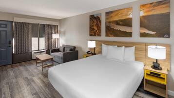 A hotel room with a large bed, a desk, a chair, and a window with curtains. There are two wall-mounted lamps and a small table.