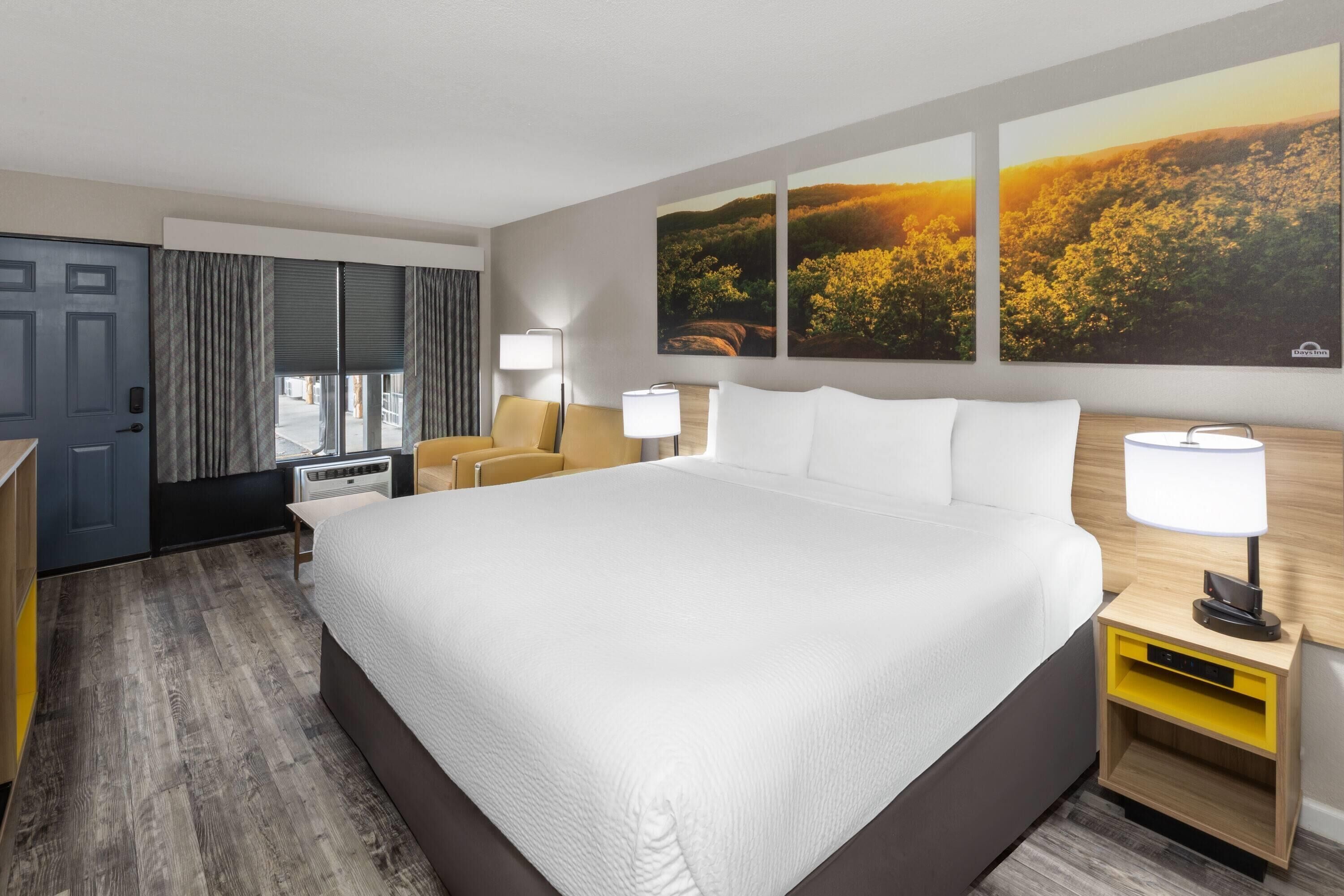 A hotel room with a large bed, a desk with a lamp, a chair, a wardrobe, and a window with curtains.
