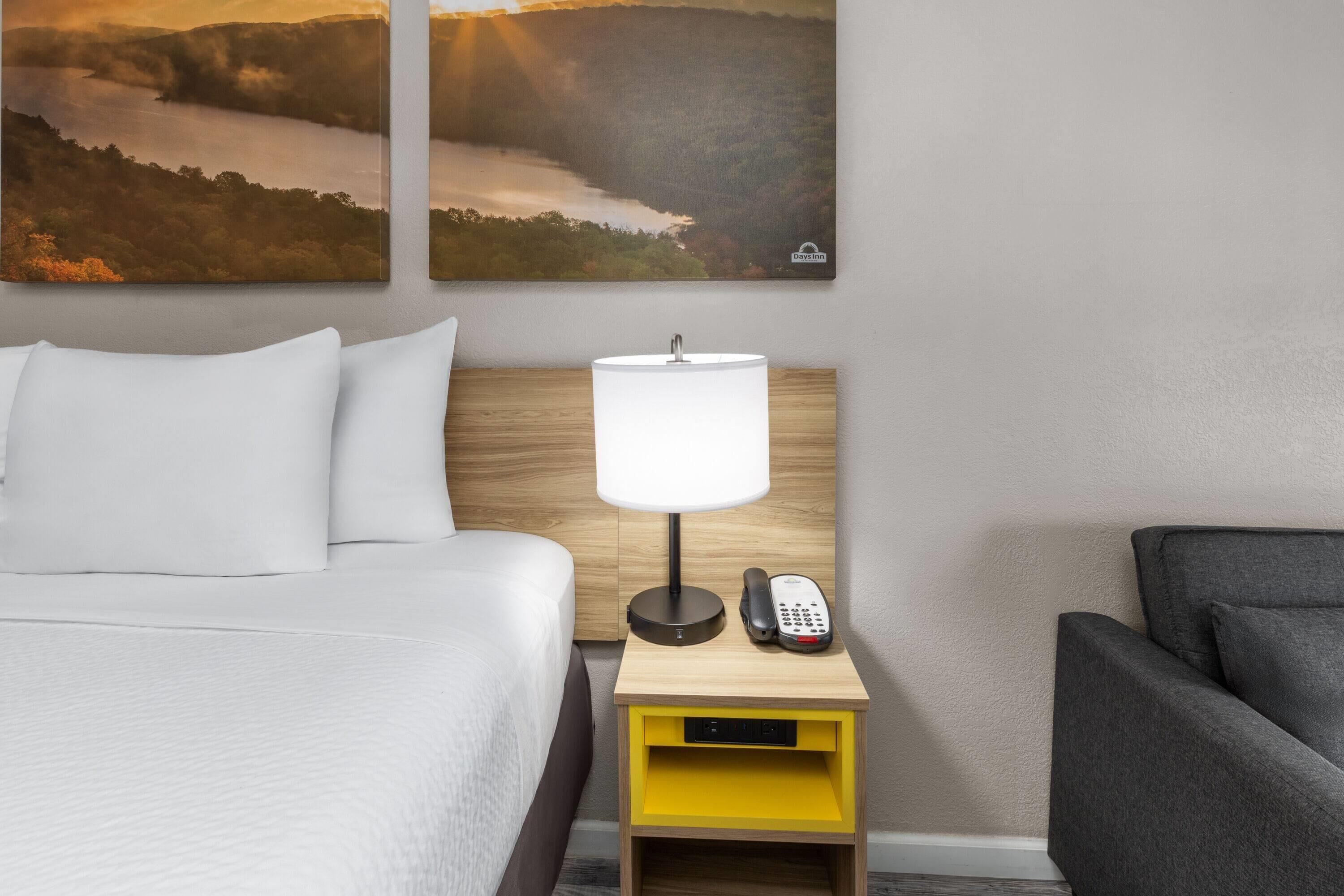 A hotel room with a bed, a nightstand with a lamp and phone, and two landscape paintings on the wall.