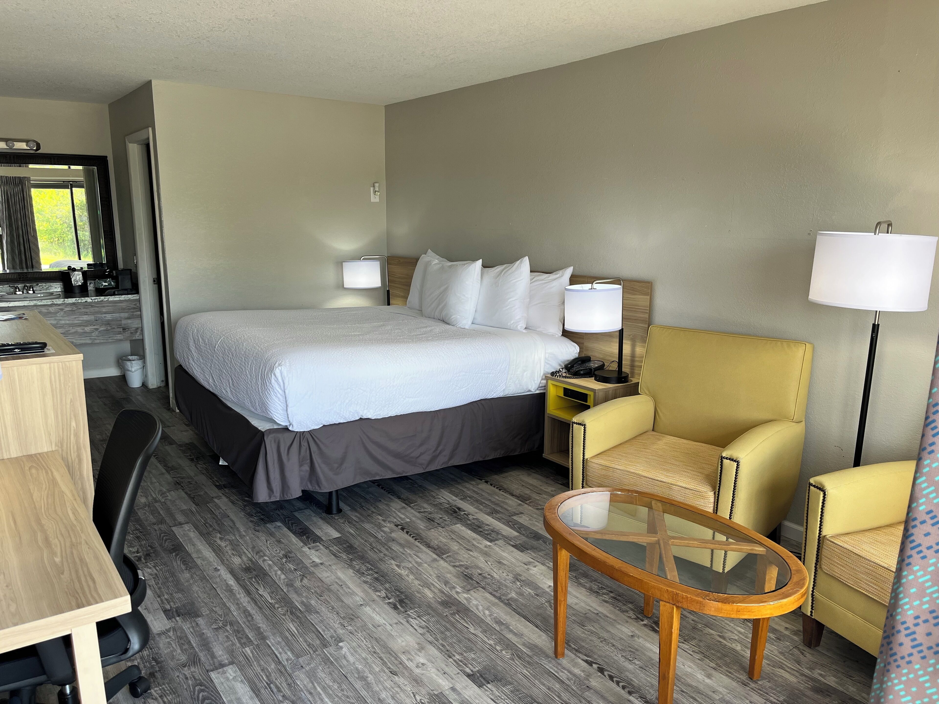 A hotel room with a large bed, a desk, a chair, a small table, and a lamp.