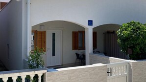 Un bâtiment moderne à la façade blanche, doté de deux portes cintrées, d’un petit espace extérieur aménagé en coin salon et d’une entrée fermée par un portail.