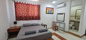 Una camera da letto con un letto, una scrivania, una sedia, un comodino, una finestra con tenda rossa, un quadro al muro e un bagno visibile attraverso una porta aperta.