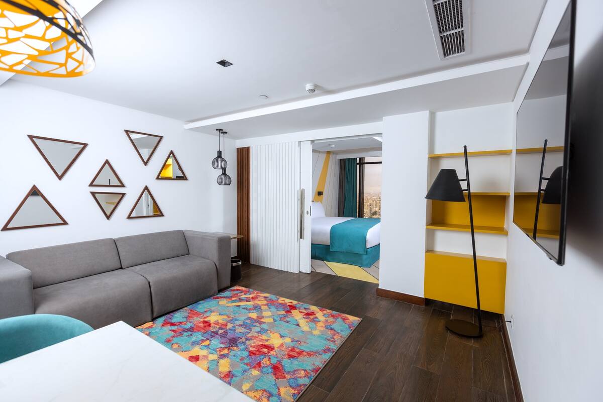 A modern hotel room with a grey sofa, colorful rug, and a bed with a blue and green bedspread.