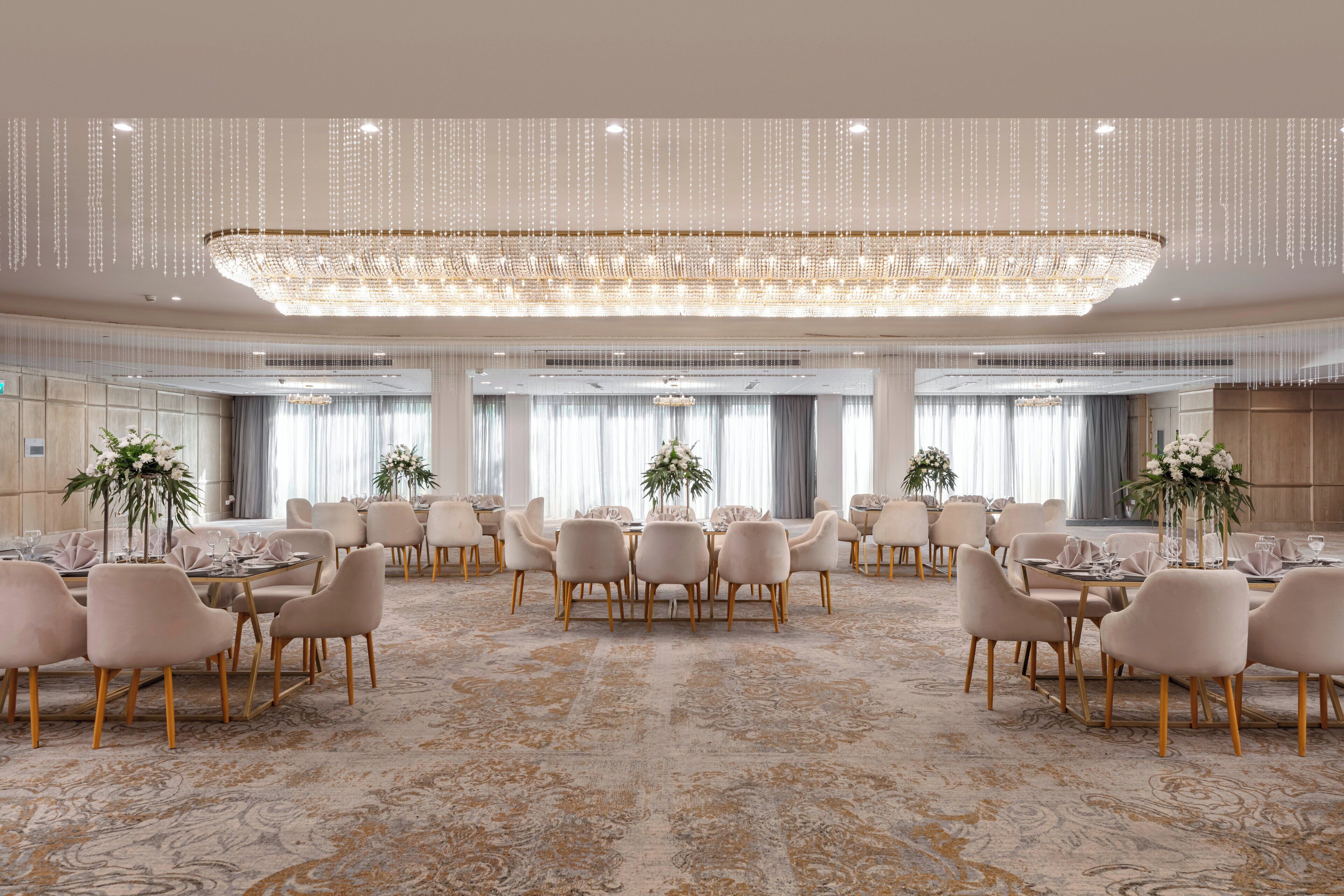A spacious banquet hall with round tables, elegant chandeliers, and a patterned carpet.