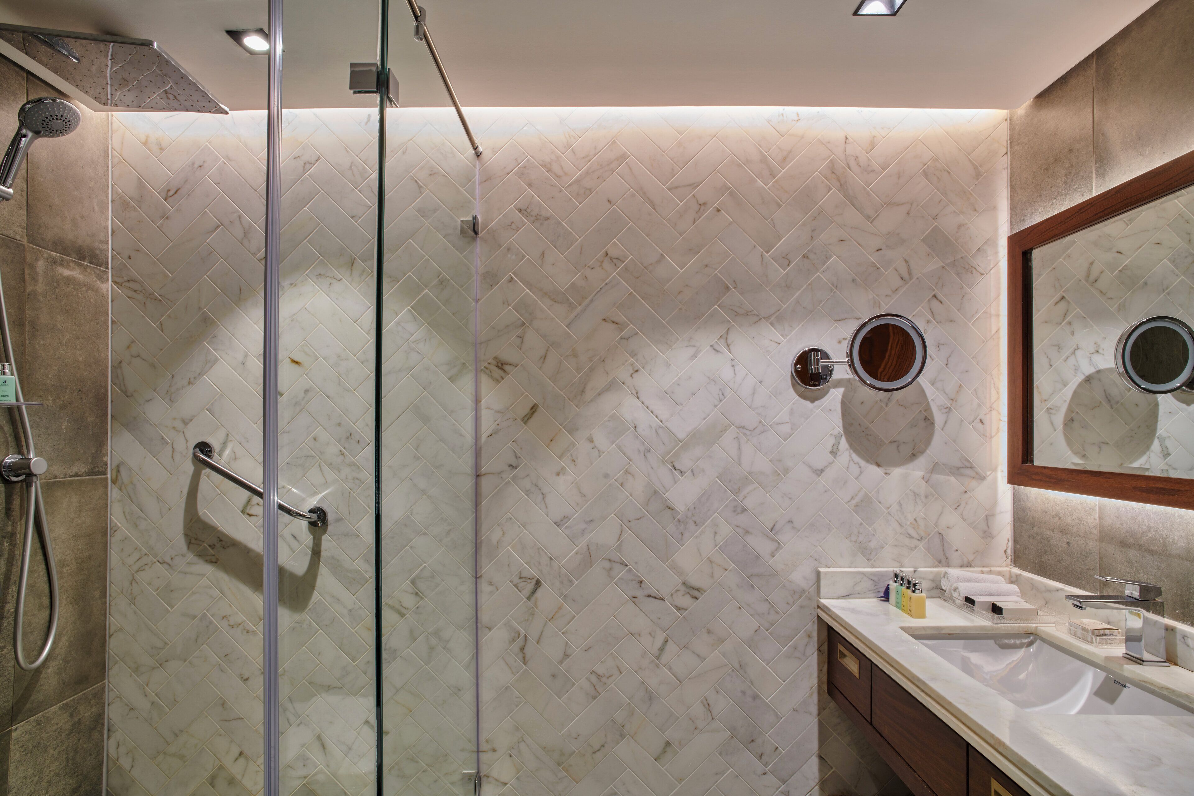 Une salle de bains moderne avec une douche à parois de verre, un mur recouvert de marbre disposé en motif chevrons, un lavabo avec un miroir et un meuble-vasque avec des articles de toilette.