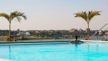 Una piscina con agua azul cristalina, rodeada de palmeras y vista a un paisaje urbano al fondo.