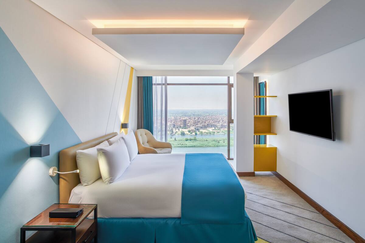 A hotel room with a large bed, a TV mounted on the wall, and a view of a cityscape through the window.