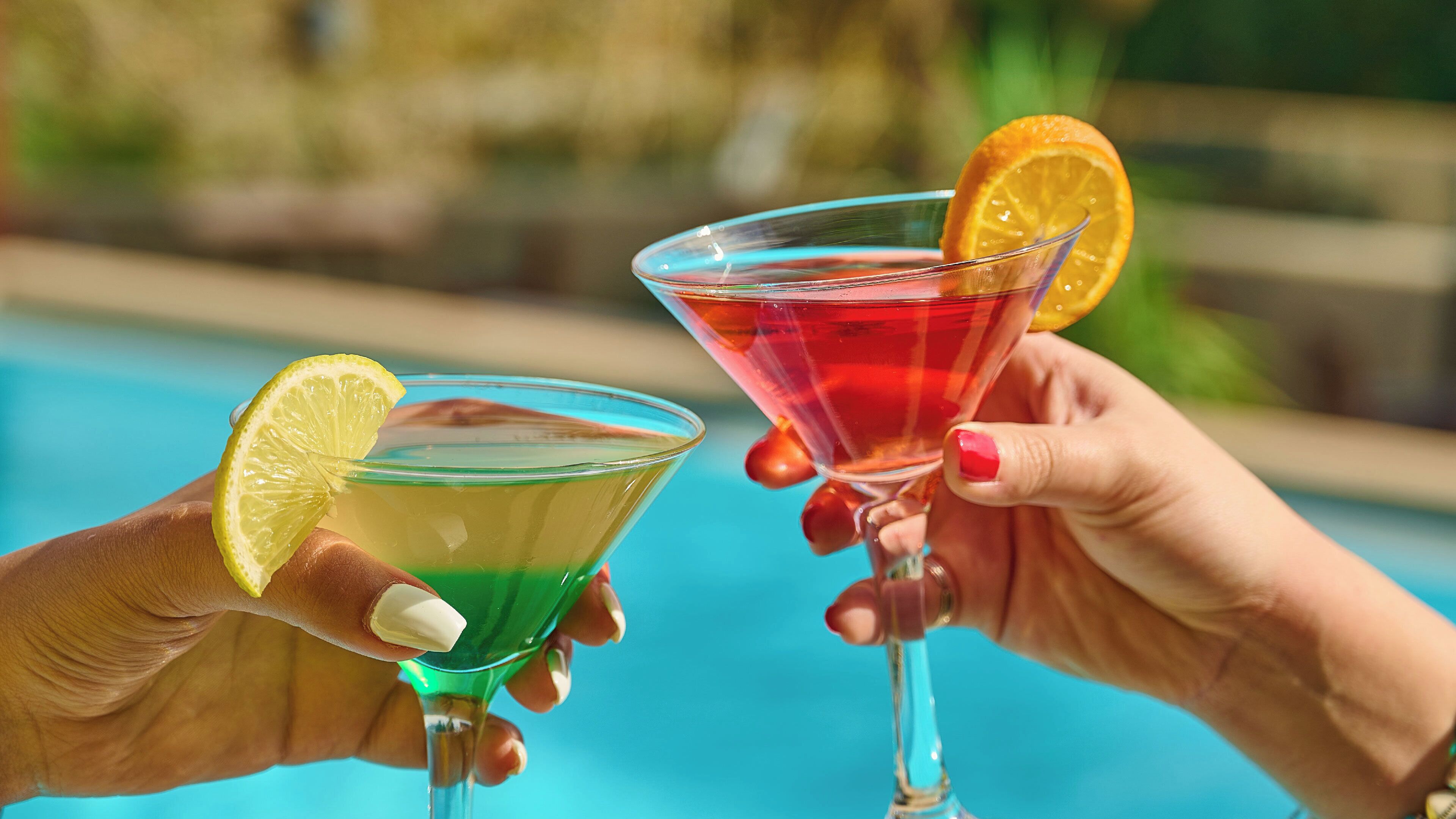 Deux mains tenant des verres à cocktail, avec une piscine en arrière-plan.