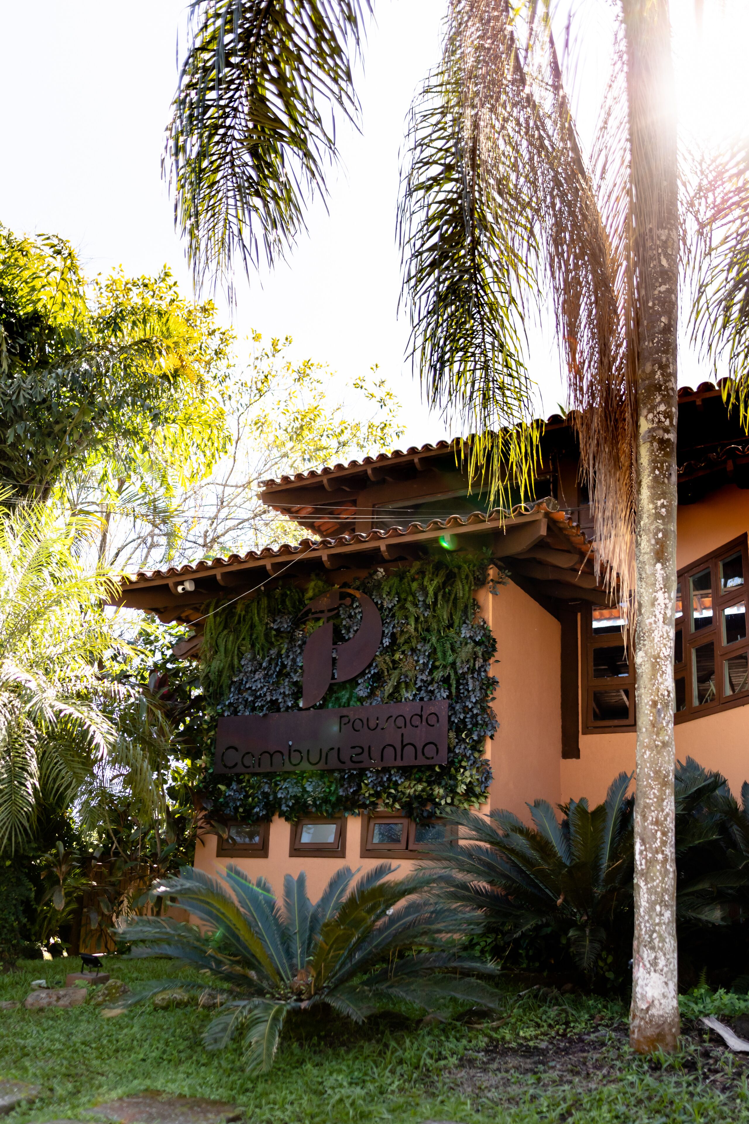 A building with a sign reading "Pouro do Camburéinha" surrounded by lush greenery and palm trees.