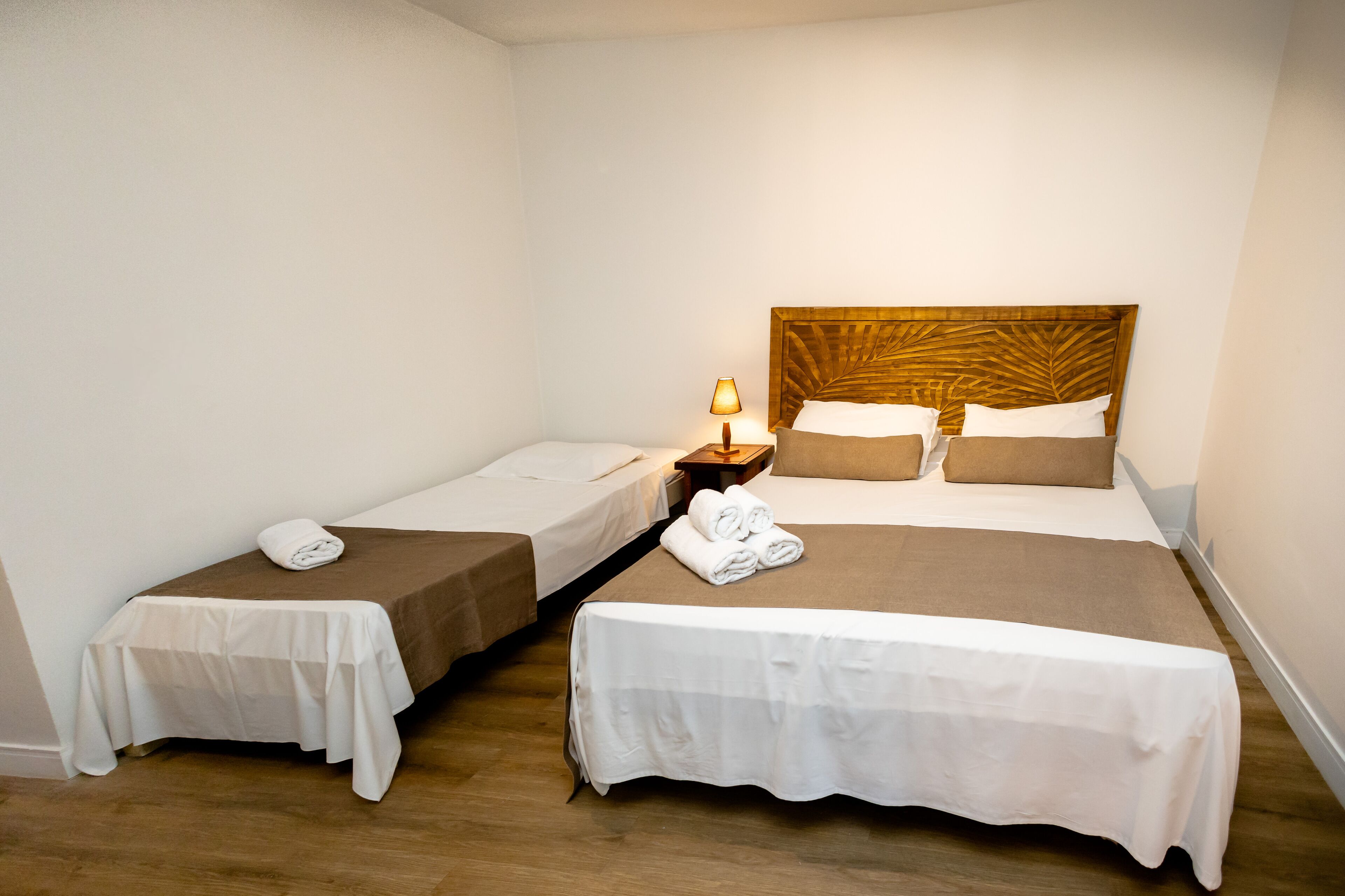 Ett hotellrum med två enkelsängar och en dubbelsäng, alla med vita sängkläder och en brun sänggavel. Det finns ett litet bord med en lampa och en hopvikt handduk på dubbelsängen.