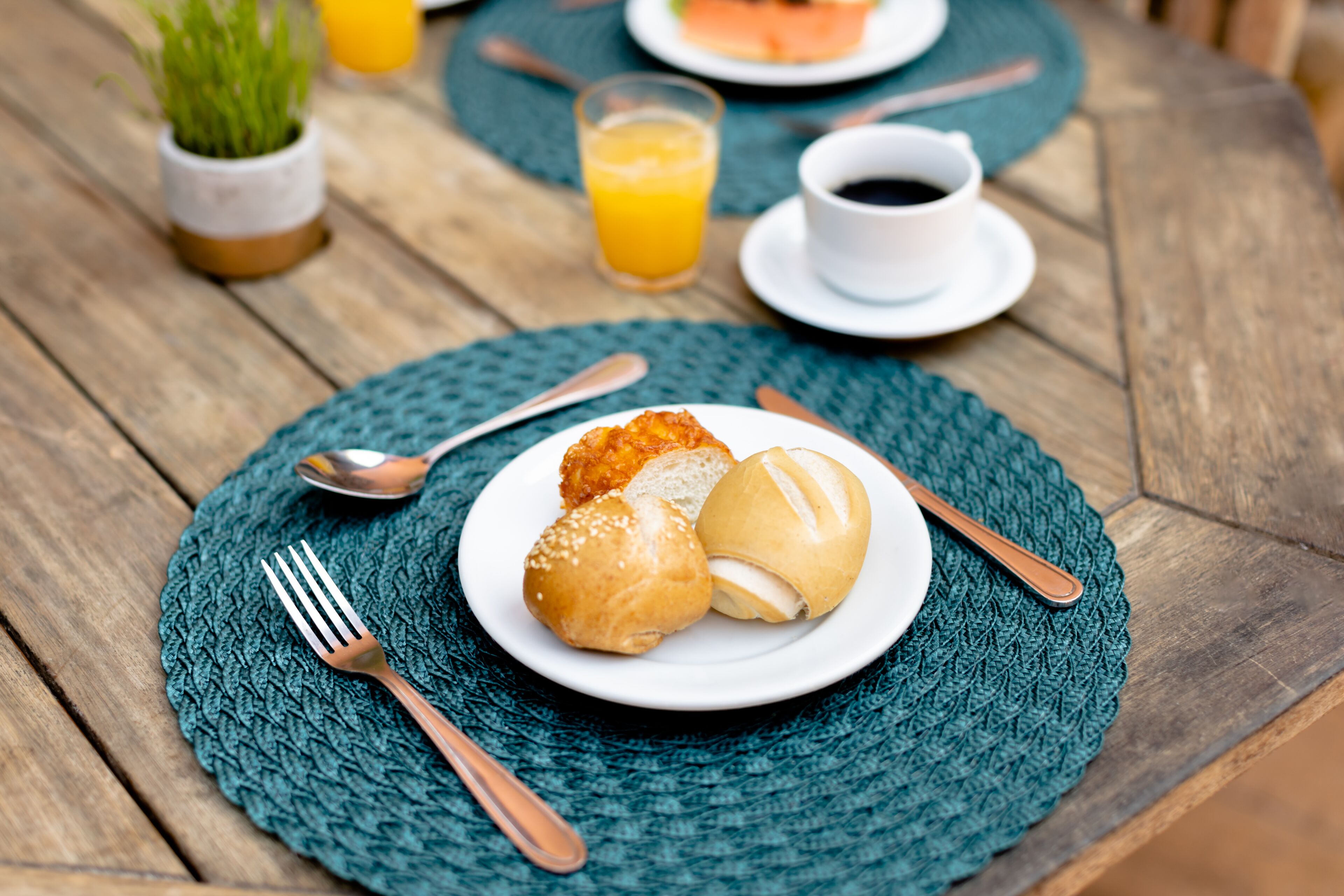 A wooden table with a woven placemat, a plate with bread rolls, a fork, and a spoon.