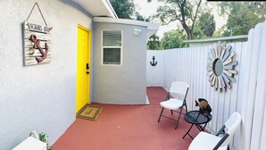 Une terrasse avec un sol rouge, des chaises blanches, une petite table et une porte jaune.