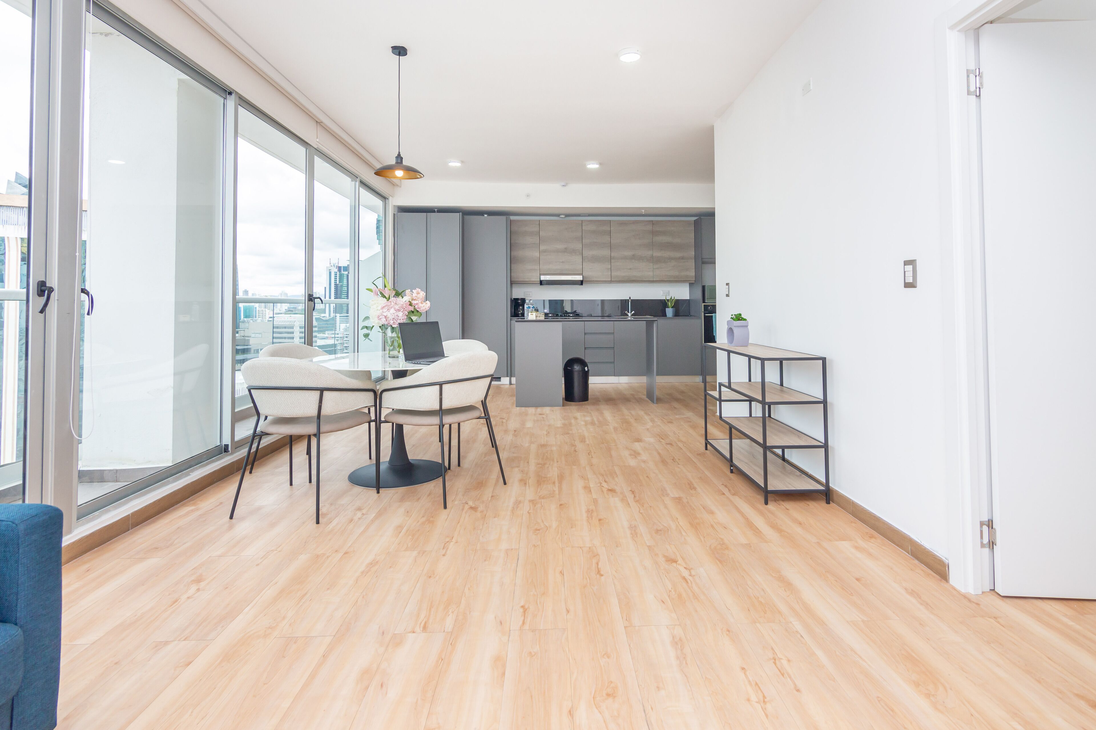 A modern apartment with a dining area, kitchen, and large windows offering a city view.