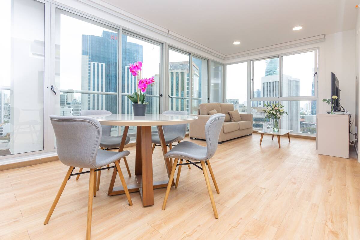 A modern living room with a round dining table, grey upholstered chairs, a beige sofa, and a small coffee table. Large windows offer a view of the cityscape.
