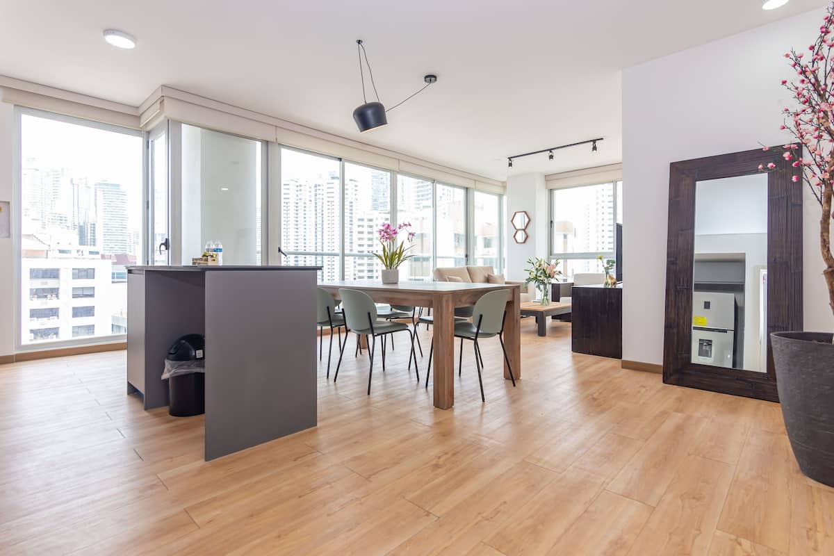 A spacious living area with a wooden floor, a dining table with chairs, a large window with a city view, and a potted plant.