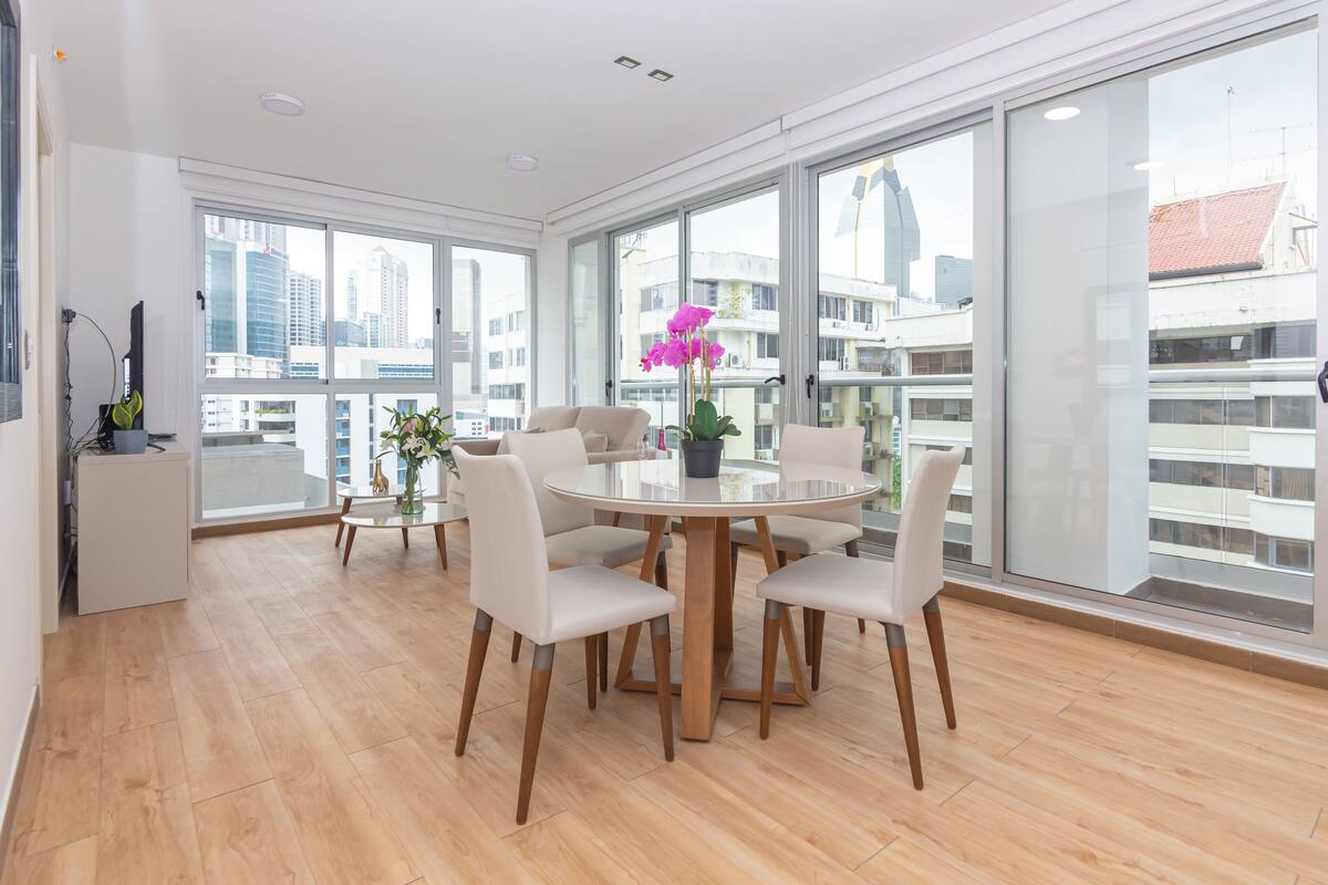A modern dining area with a round table and chairs, large windows with city views, and a potted orchid.
