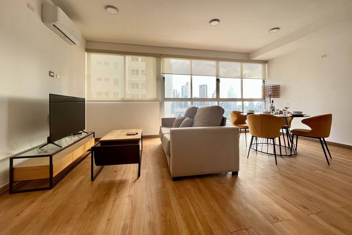 A modern living room with a flat-screen TV, a sofa, a dining table with chairs, and large windows offering a city view.