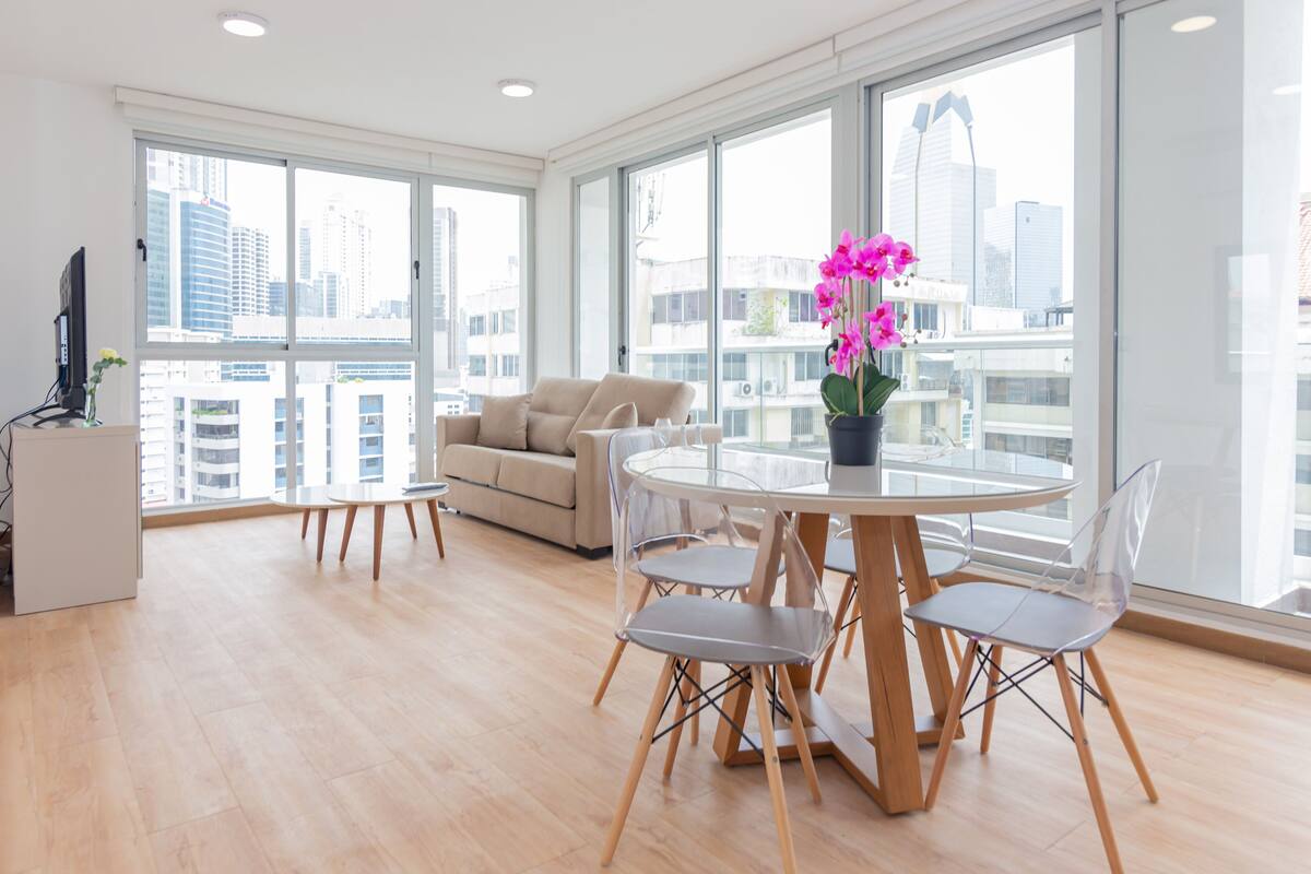 A modern living room with a round dining table, clear chairs, a sofa, and a large window offering a city view.