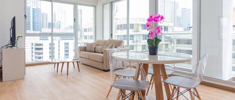 A modern living room with a round dining table, clear chairs, a sofa, and a large window offering a city view.