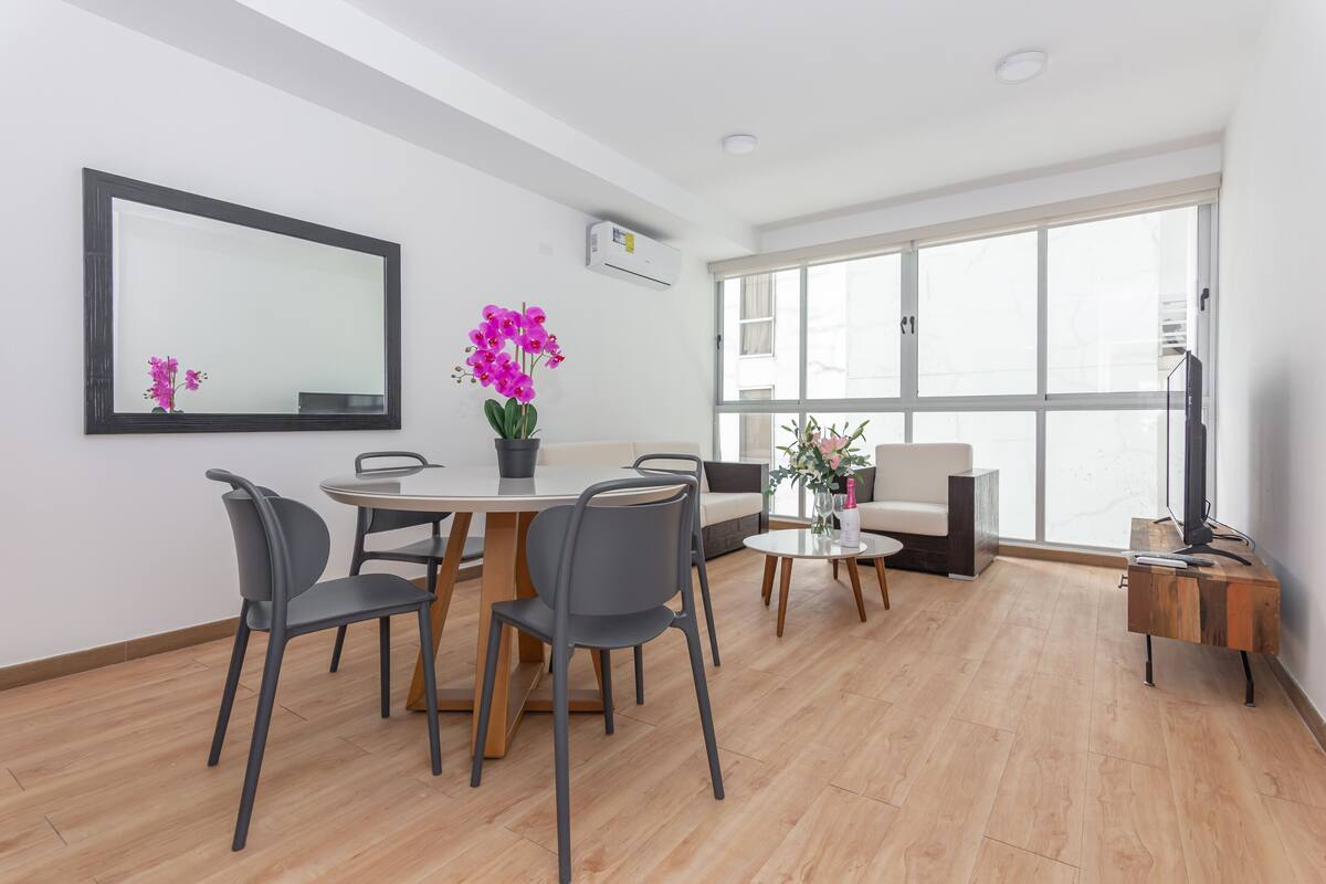 A modern living room with a dining area, large windows, and a flat-screen TV.