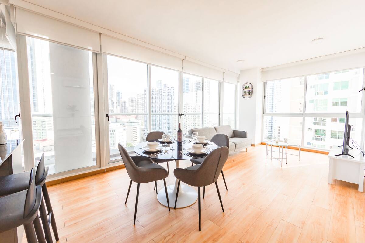 A modern dining area with a round table set for four, large windows offering a city view, and a white desk with a computer.