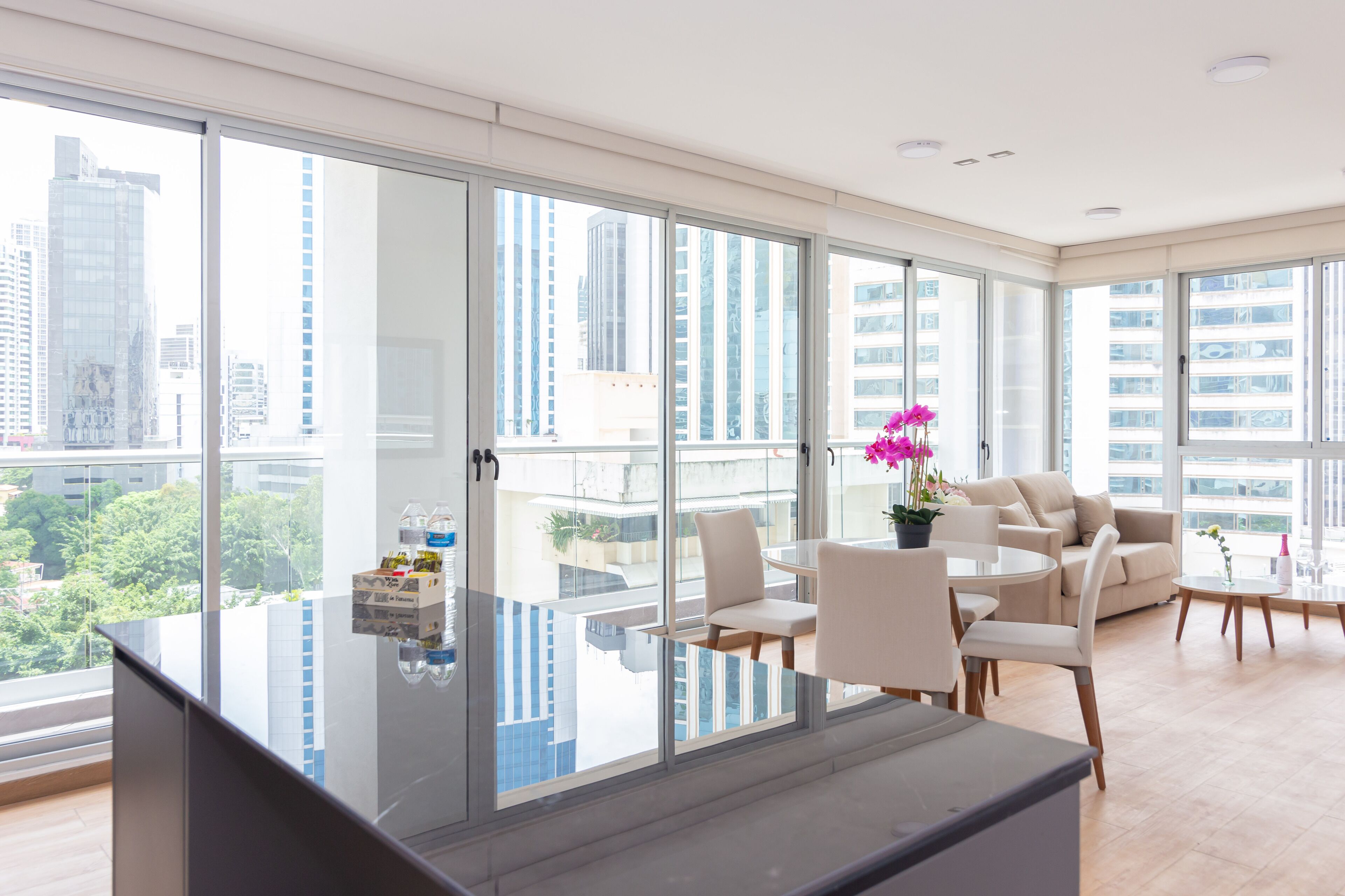 A modern living room with a glass partition, a kitchen area, and a dining space with a view of the cityscape.