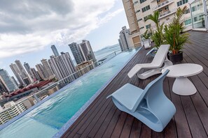 Zona de piscina en la azotea con sillones y una mesa, con vistas a un paisaje urbano de edificios altos.