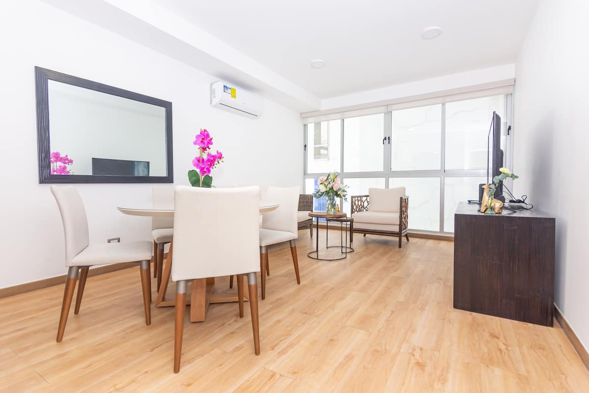 A modern living room with a dining area, large windows, a flat-screen TV, and a dark wooden side table.