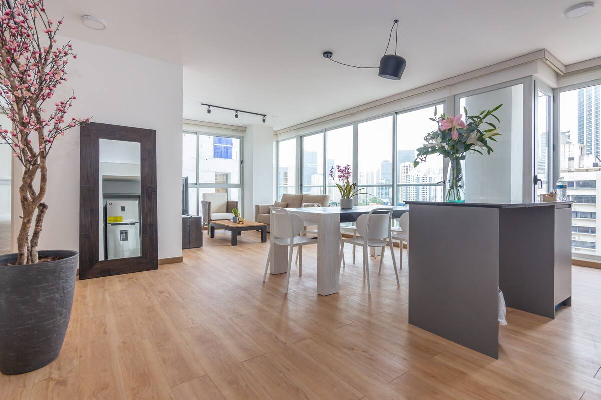A spacious living area with a dining table and chairs, a large potted plant, and a view of the city through large windows.