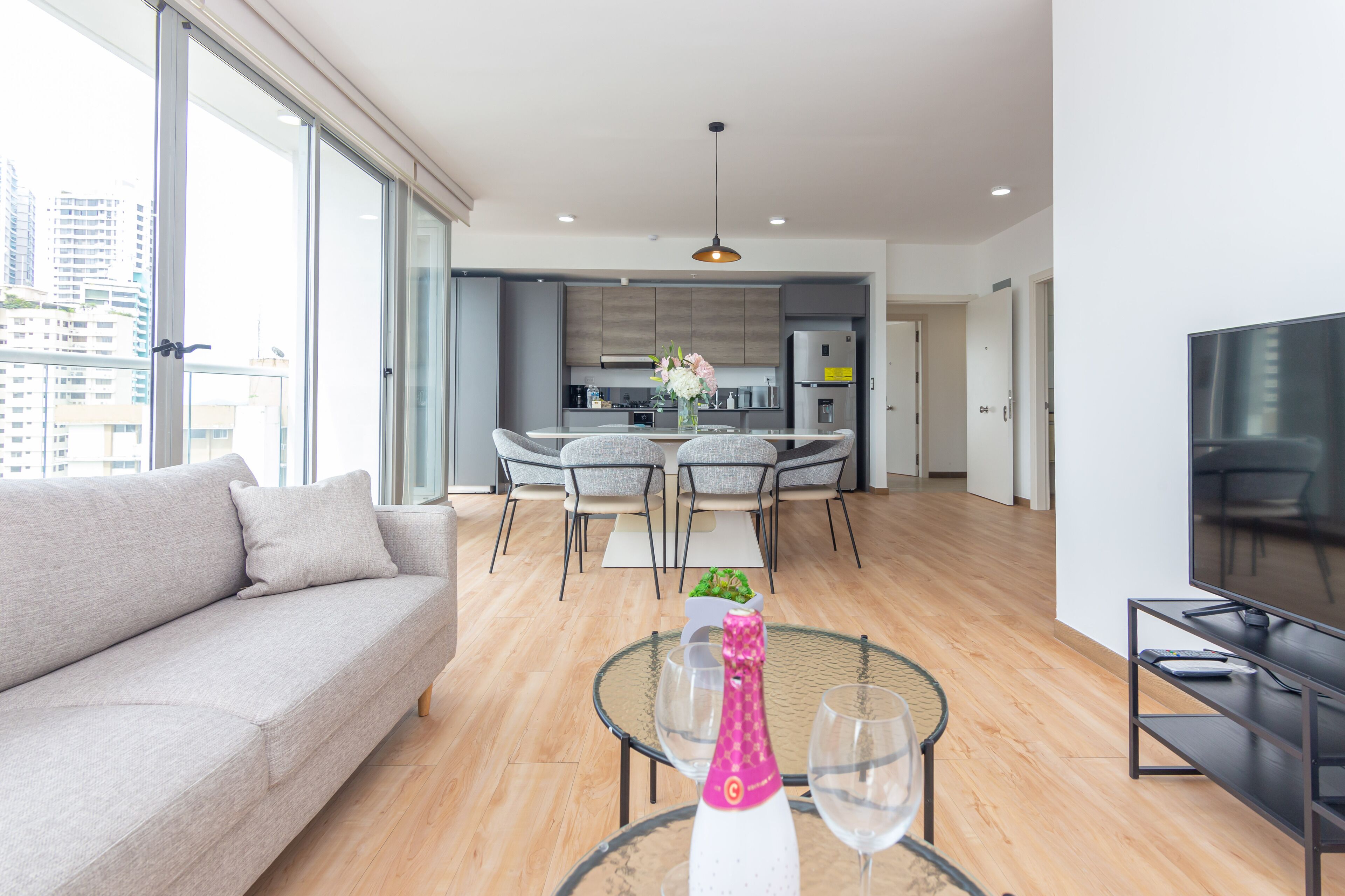 A modern living room with a sofa, a dining table with chairs, a kitchen in the background, and a TV on a stand.