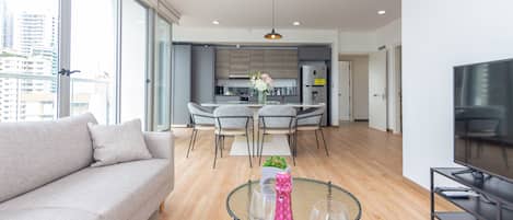A modern living room with a sofa, a dining table with chairs, a kitchen in the background, and a TV on a stand.