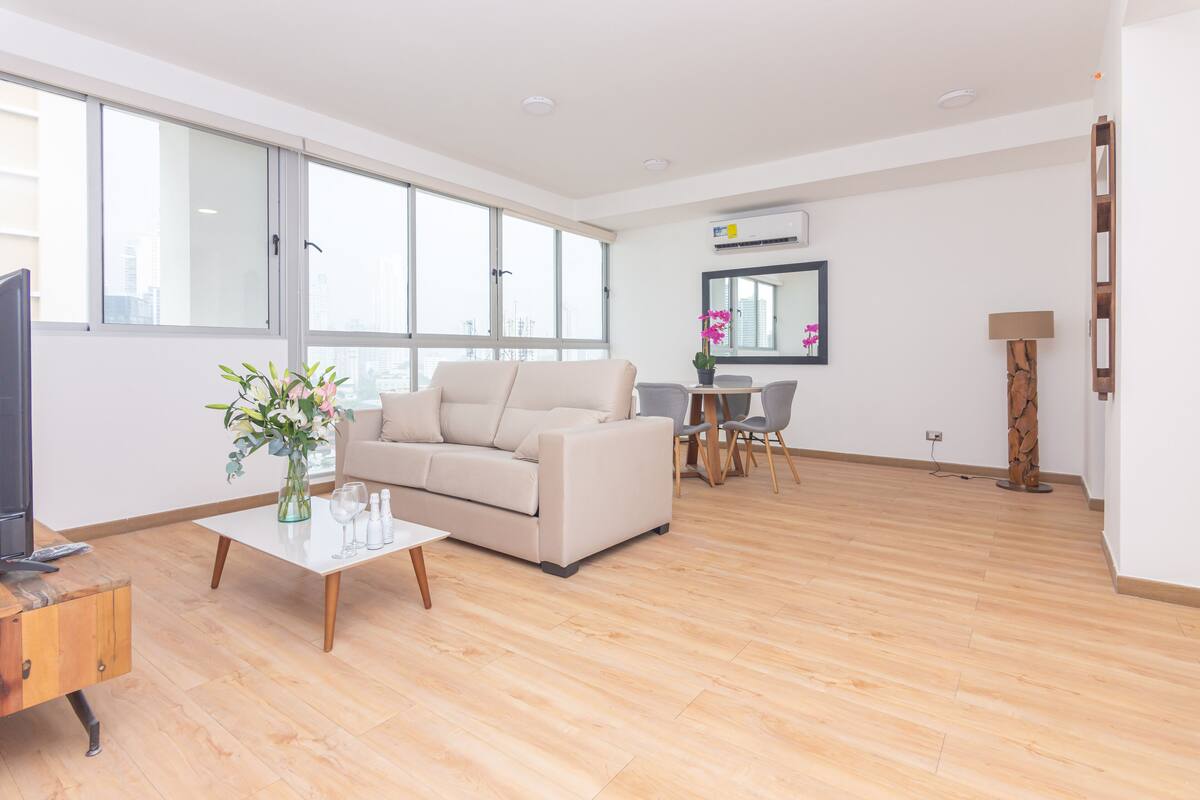 A modern living room with a sofa, coffee table, and a TV. There is a dining area with chairs and a table. Large windows provide natural light.