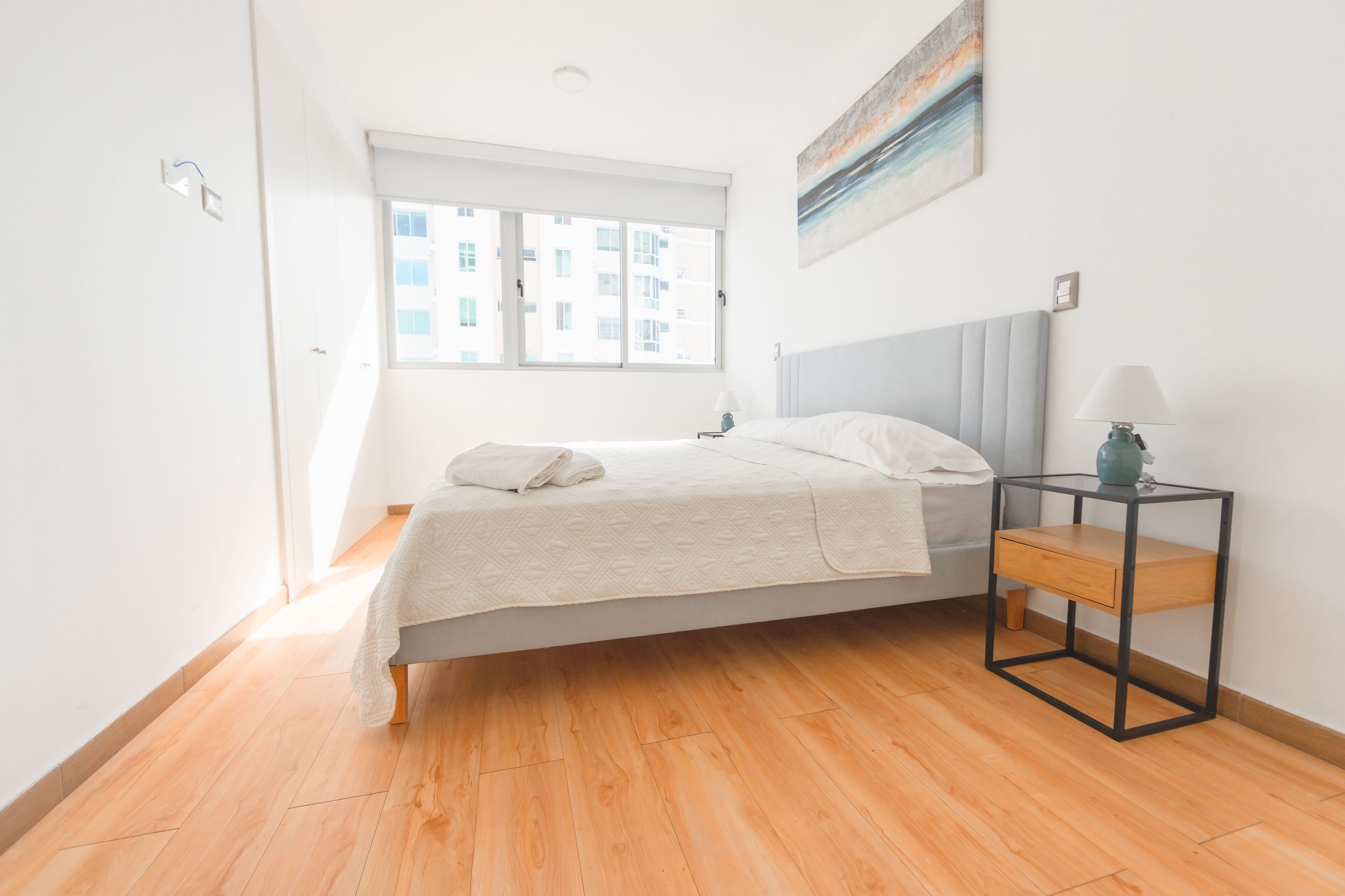 Un dormitorio con cama, mesita de noche, ventana y un cuadro en la pared.
