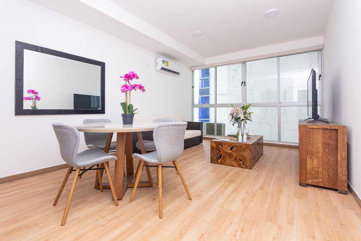 A modern living room with a dining table, two grey chairs, a wooden coffee table, and a large window with a view.