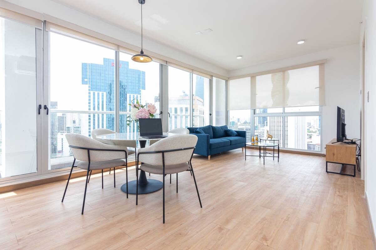 A spacious living area with a large window, a blue sofa, a dining table with chairs, and a TV stand.