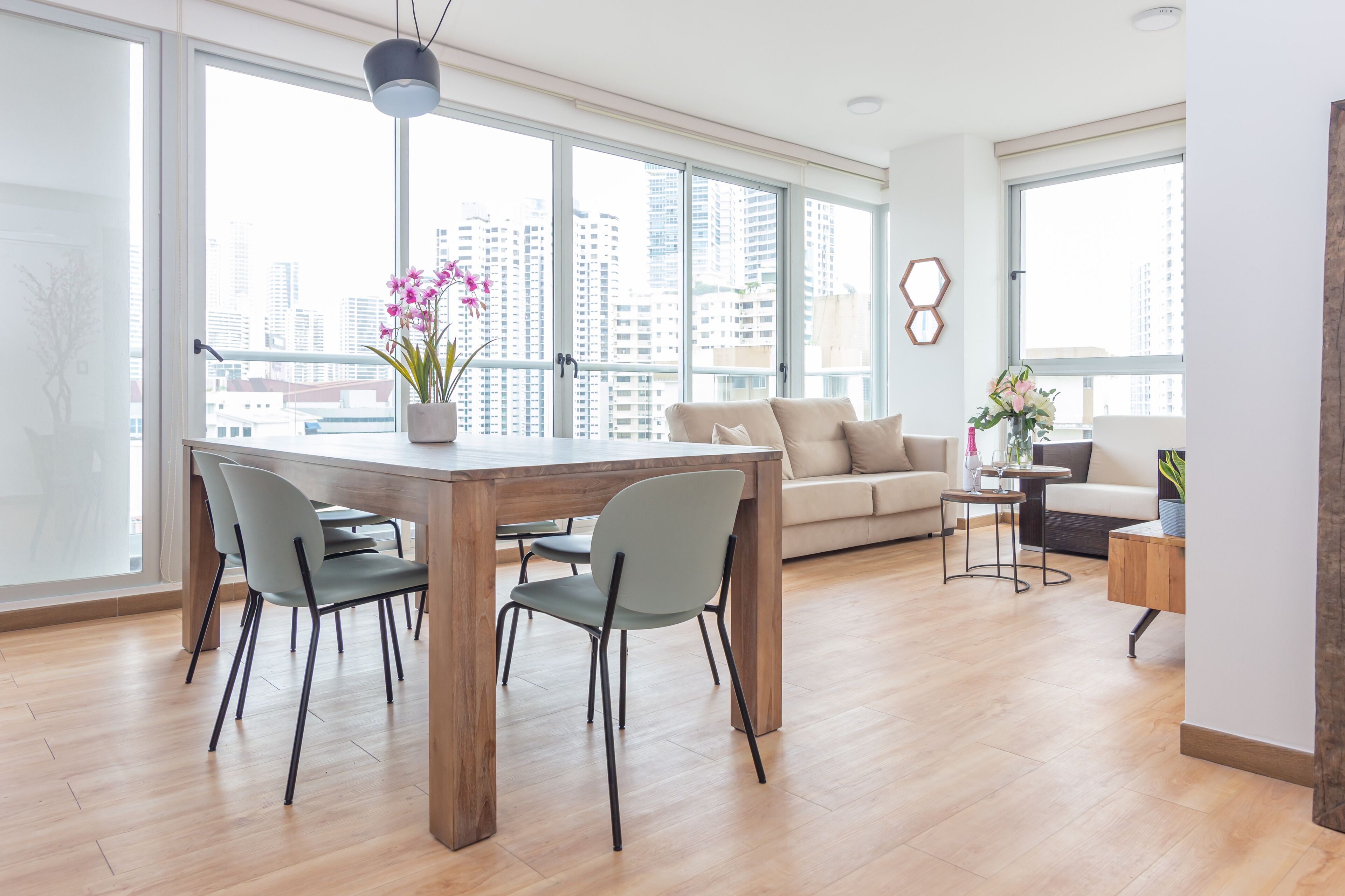 Una sala de estar moderna con una mesa de comedor de madera, sillas grises, un sofá color beige y vistas a la ciudad a través de amplios ventanales.