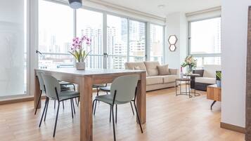 A modern living room with a wooden dining table, grey chairs, a beige sofa, and a city view through large windows.