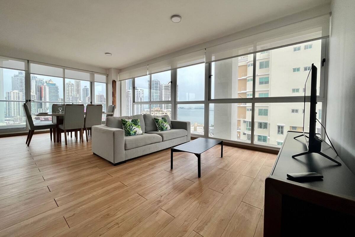 A spacious living room with a sofa, coffee table, and dining area. Large windows offer a view of the cityscape.