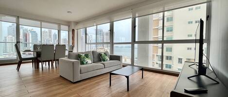 Amplia sala de estar con sofá, mesa de centro y comedor. Ventanas grandes ofrecen vistas del paisaje urbano.