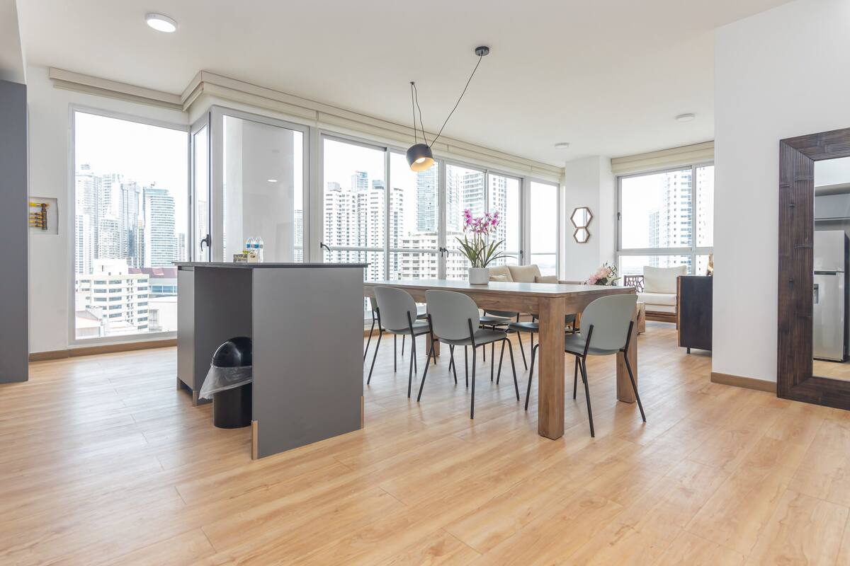 A spacious living area with a dining table, chairs, and a large window offering a city view.